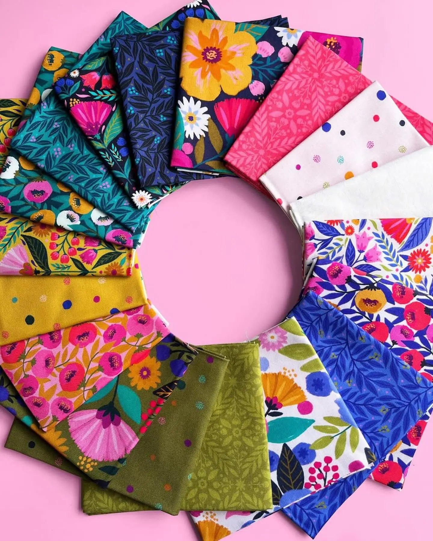 A circular display of the Dayspring Fat Quarter Bundle by Windham Fabrics shows vibrant floral, geometric, and polka dot quilting fabrics in blue, pink, green, yellow, and white on a pink background.