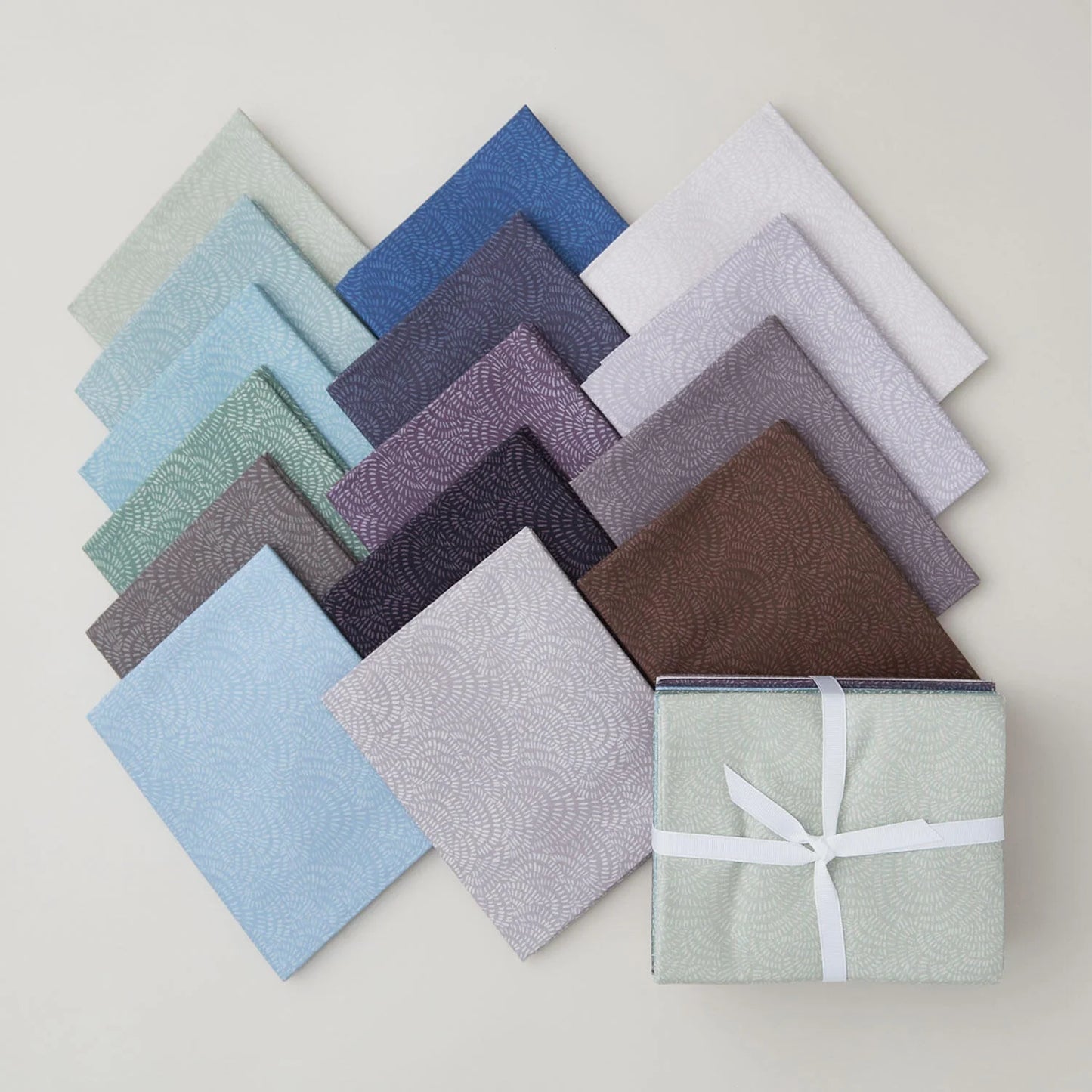 A selection of neatly arranged fabric squares in various cool colors and subtle patterns, including the Crimp Cool Colors Fat Quarter Bundle by Dear Stella, with one bundle tied with a white ribbon, displayed on a light surface.