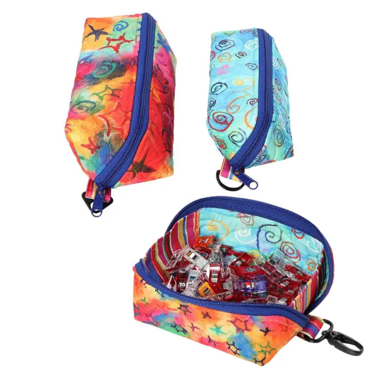 Three small, colorful Drop Zone pouches by Quilted Joy feature blue zippers and swirl/star patterns; one is open showing clear clips inside, while the other two are closed and empty.
