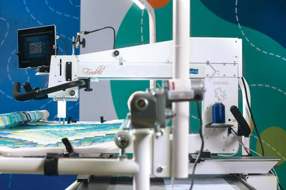 The APQS Demo Freddie 26" Long Arm Quilting Machine is stitching a vibrant geometric quilt. Featuring a mounted screen and multiple control handles, it's perfect for display demos against an abstract background.
