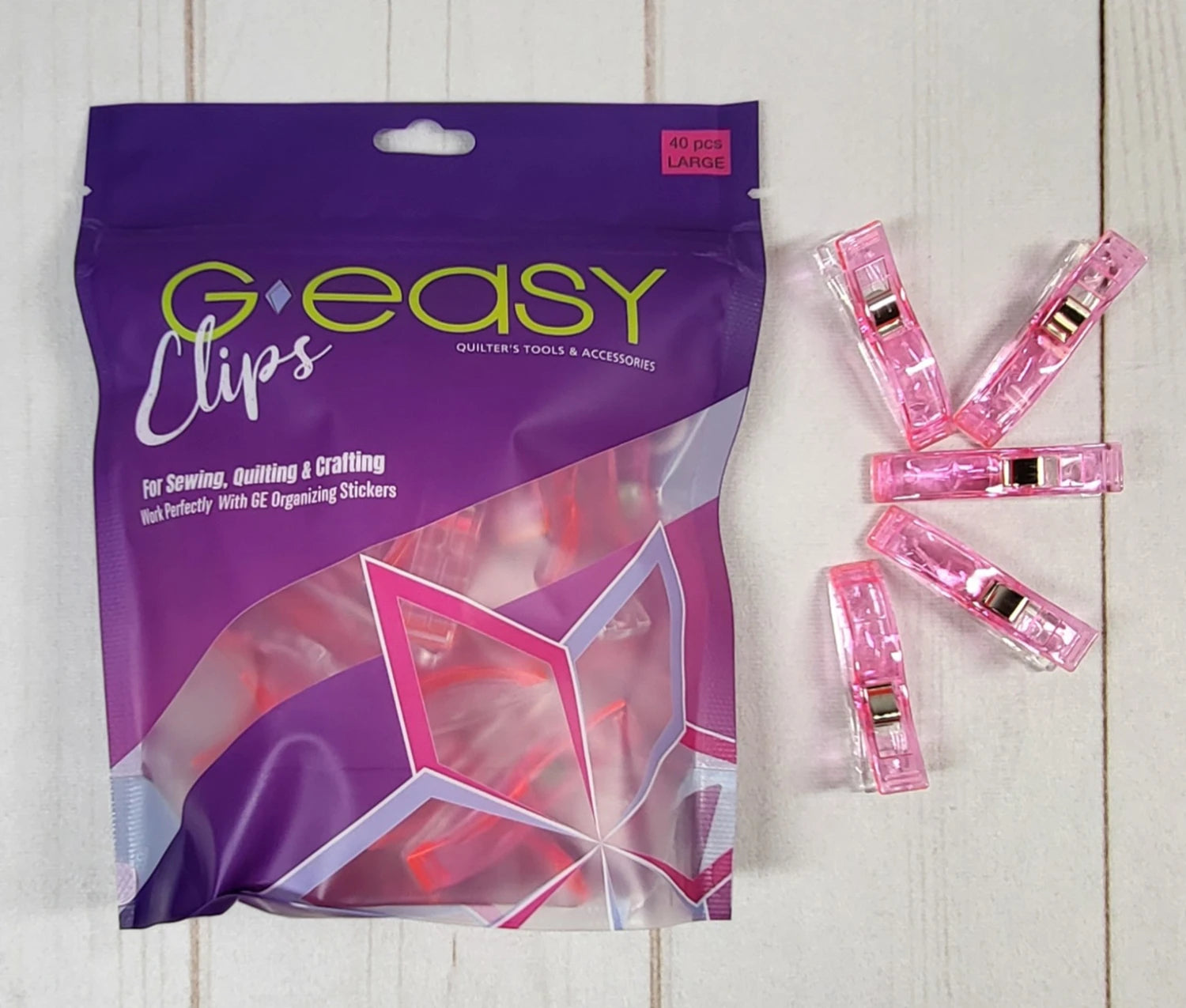 A package of Checker Distributor’s GEasy Large Pink Organizing Clips (40-count) sits on a light wooden surface, with six pink clips arranged beside the bag. These clips are ideal for sewing and crafting projects.