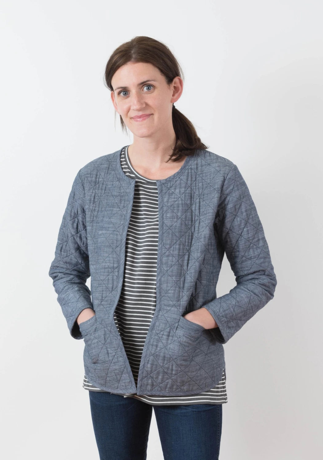 A woman with brown hair in a ponytail wears the Checker Distributor Tamarack Jacket (Sizes 0-18) over a black-and-white striped shirt and dark jeans, standing against a white background—ideal for spring and fall layering.