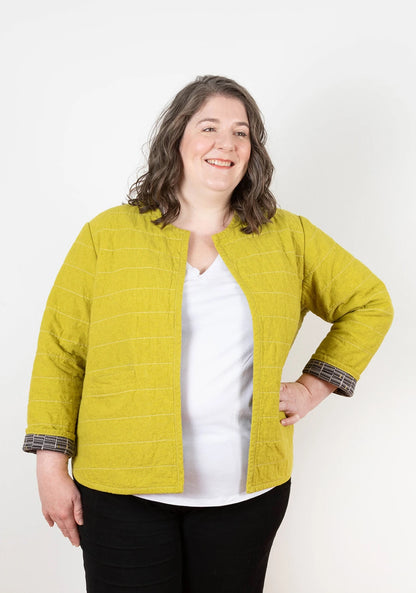 A woman with wavy, shoulder-length brown hair smiles while wearing a mustard yellow Checker Distributor Tamarack Jacket (Sizes 14-30) over a white shirt and black pants, standing against a plain white background.