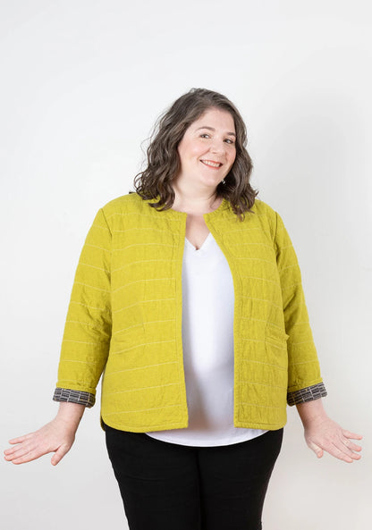 A smiling woman with wavy brown hair wears the Checker Distributor Tamarack Jacket Sizes 14-30 in bright yellow over a white top and black pants, standing against a plain white background with her hands slightly out to the sides.