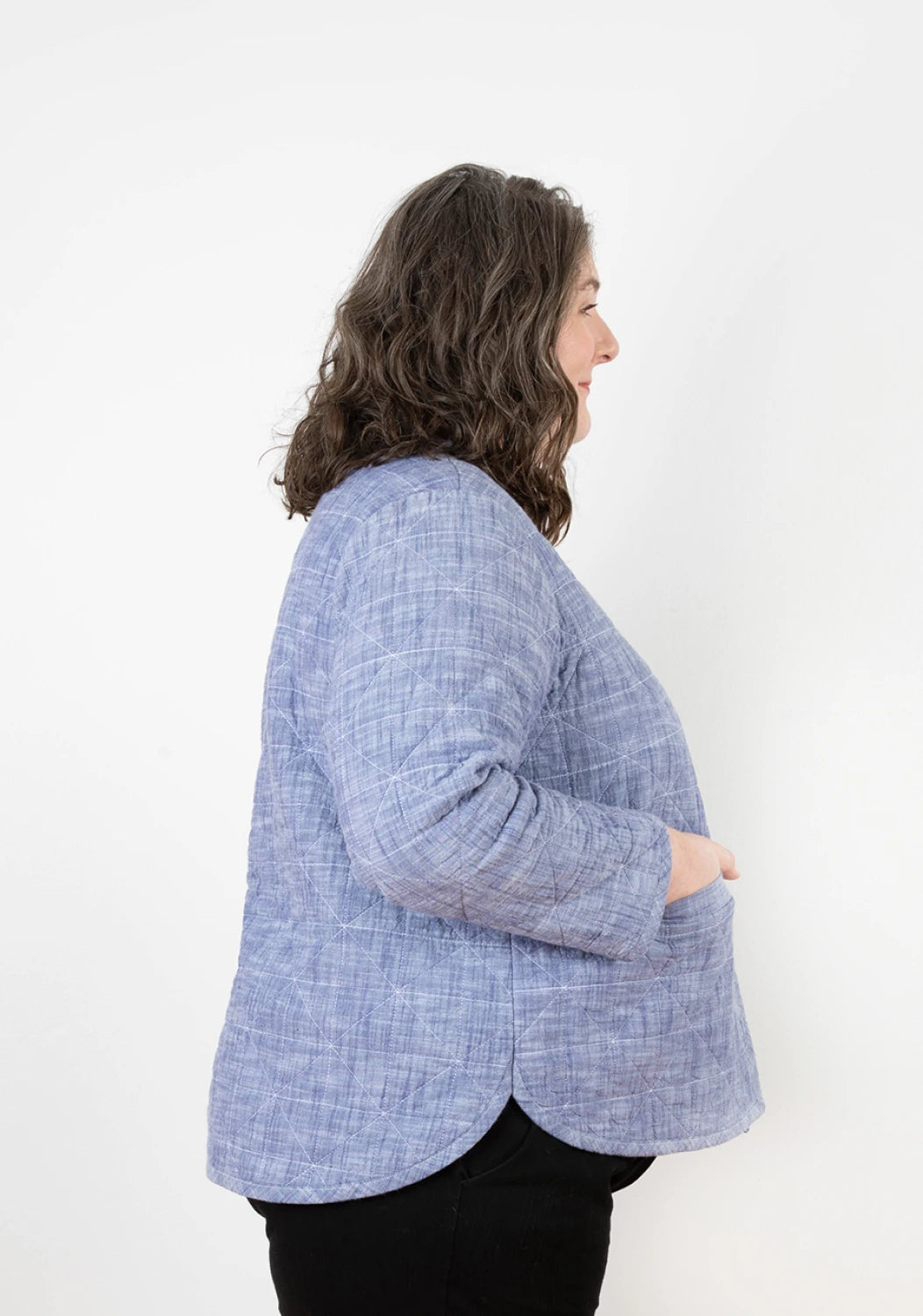 A woman with wavy brown hair models the Checker Distributor Tamarack Jacket Sizes 14-30 in textured light blue, paired with black pants, standing in profile against a plain white background with her hands in her jacket pockets.