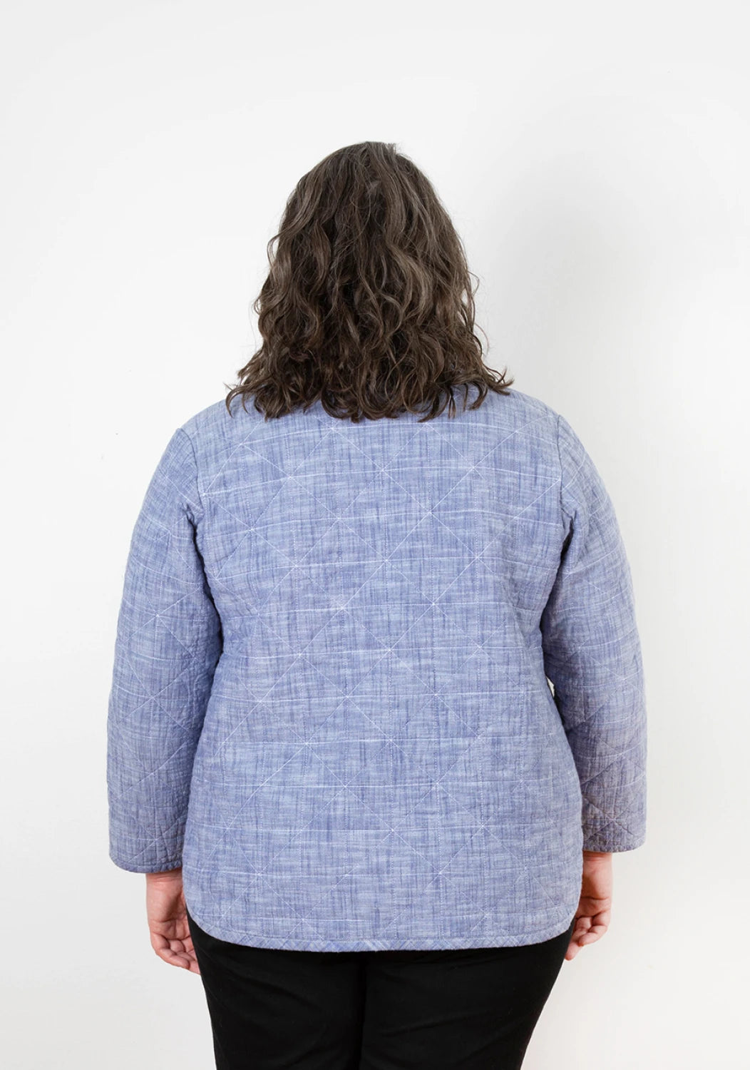 A person with wavy brown hair is seen from behind, wearing a light blue, quilted coat made with the Checker Distributor Tamarack Jacket Sizes 14-30 pattern and black pants, standing against a plain white background.