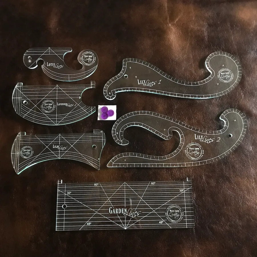 The White Arbor Quilting Garden Lines Collection Ruler Set includes five transparent acrylic French curve templates, a rectangular ruler with white markings, and four purple circular grips, all arranged on a brown leather surface.