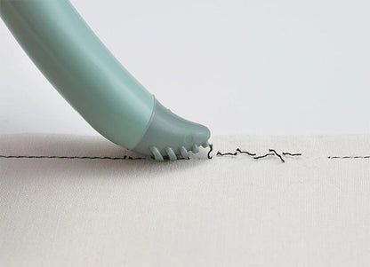 A close-up of the Grip Fit Seam Ripper, a light green handheld device with an ergonomic handle and rounded tip making a wavy black stitched line on white fabric. Small protrusions at the tip gently touch the fabric.