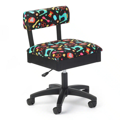 The Arrow Hydraulic Black Sewing Notions Chair features a cushioned seat and backrest upholstered in fabric with colorful sewing-themed patterns, such as scissors, pins, and thread, on a black background.
