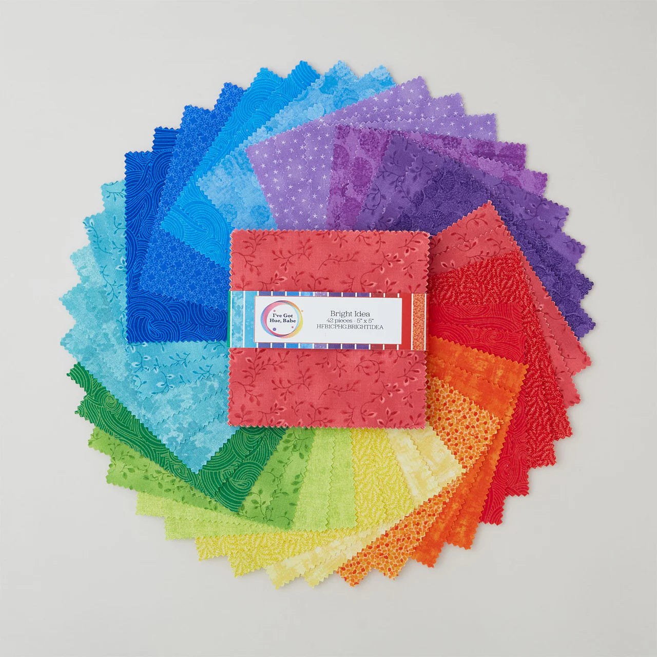 A circular rainbow gradient of colorful 5" I've Got Hue, Babe Bright Idea squares by Jaftex - Henry Glass Fabrics surrounds a neatly labeled charm pack at the center, perfectly showcasing these quilt precuts on a white background.