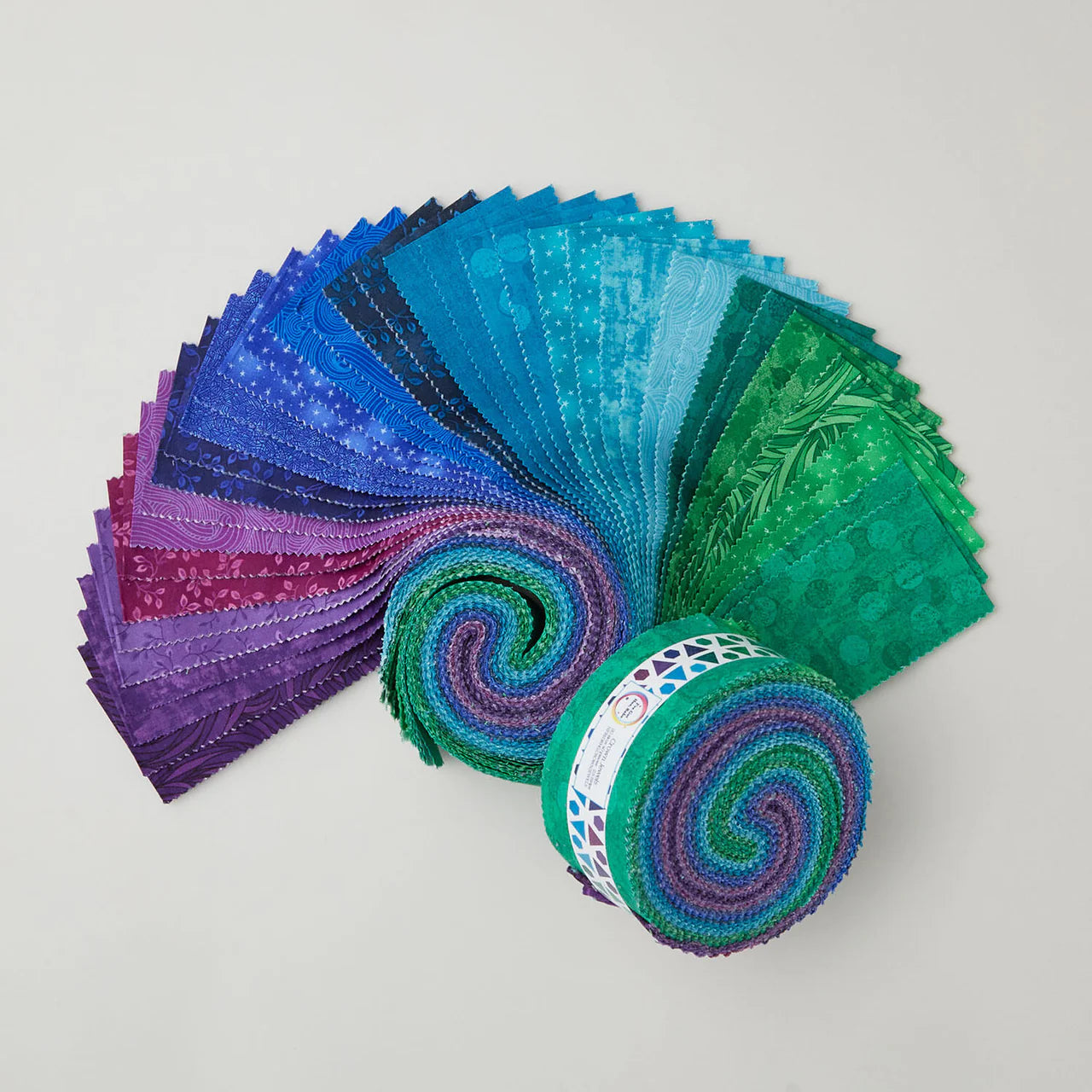 The Jaftex - Henry Glass Fabrics "I've Got Hue, Babe Crown Jewels 2.5in Strips" features blue, green, aqua, and purple tones displayed in a partially unrolled fan shape on a plain light background to show the gradient of patterns.