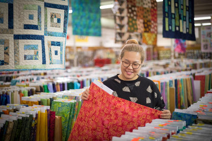 A person with glasses admires vibrant quilts and colorful quilting supplies under a bolt of red fabric, celebrating the Bus To Paducah 2026 by Quilted Joy.