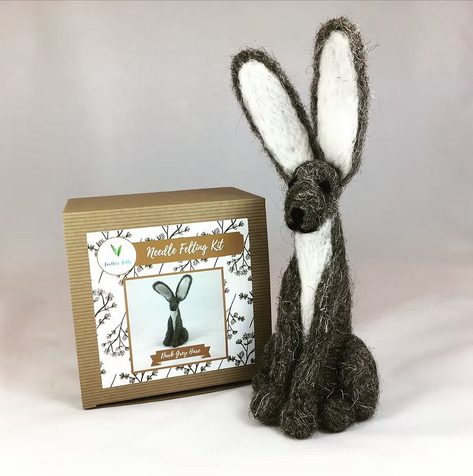 A handcrafted hare needle felting figure sits next to the Feather Felts Grey Hare - Needle Felting Kit No Foam, which features an illustration of the same hare on its box, both set against a plain, light background.