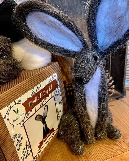 A handmade grey hare with white accents sits next to the Feather Felts Grey Hare - Needle Felting Kit No Foam box, which features its image. Wool materials are in the background, making this kit ideal for beginner needle felting.