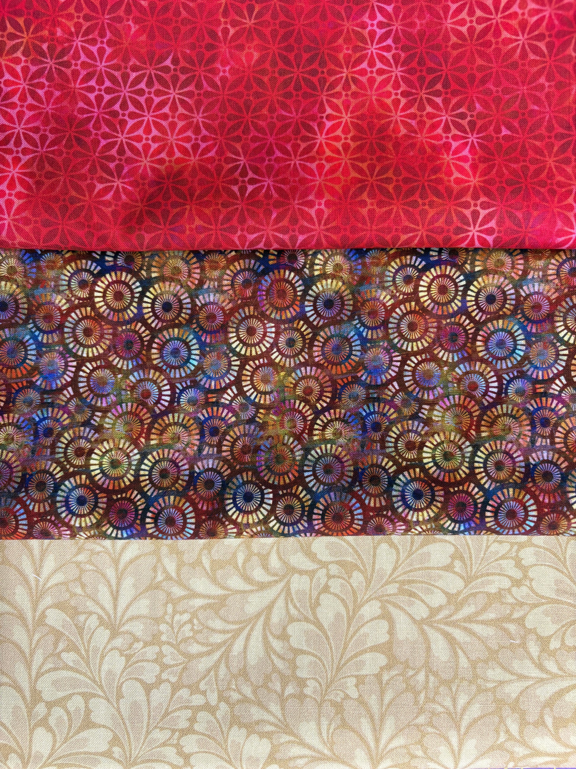 The Gilded Halcyon 3 Yard Quilt Top Bundle by Quilted Joy features a red geometric starburst, a multicolored circular print on dark, and a beige leafy swirl—perfect for pairing with Fabric Cafe 3-yard quilt books. Ideal shop-quality fabric!.