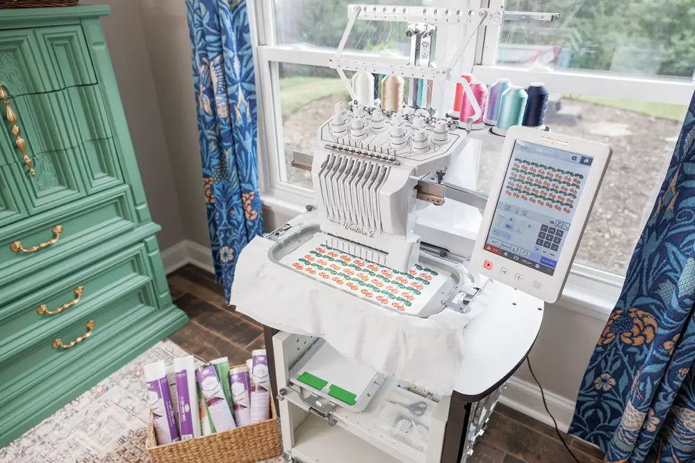 A Baby Lock Venture 2 Multi Needle Embroidery Machine by Baby Lock is embroidering colorful designs on white fabric near blue floral curtains, surrounded by spools of thread and a green cabinet—perfect for any embroidery business.