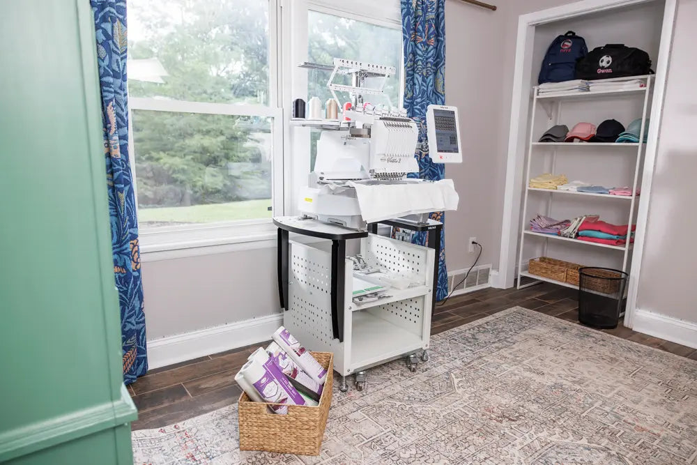 A Baby Lock Venture 2 Multi Needle Embroidery Machine by Baby Lock sits on a rolling cart near a window in a bright room, with a basket of thread stabilizer on the floor and shelves of folded clothes and bags against the wall.