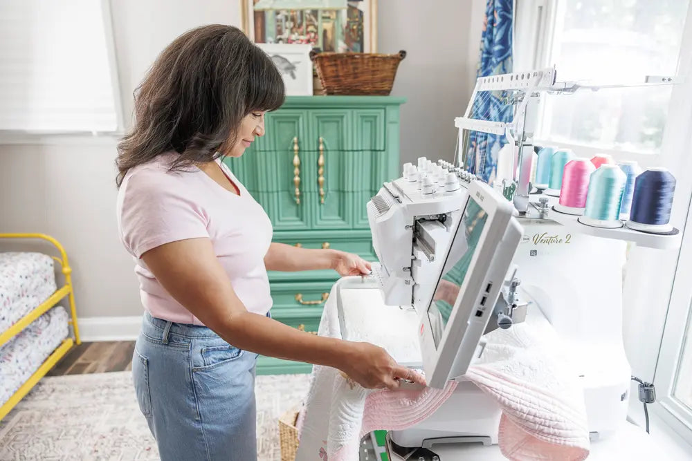 A woman uses a Baby Lock Venture 2 Multi Needle Embroidery Machine by Baby Lock in a bright room. Colorful thread spools top the machine, with a green cabinet and bed enhancing her vibrant embroidery business space.
