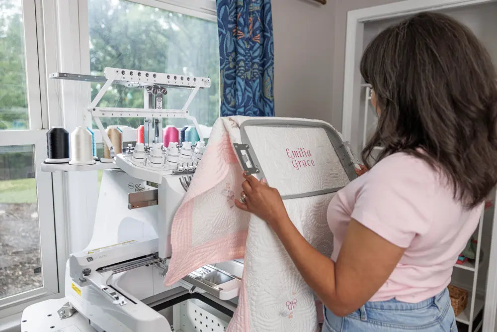 A woman stands by her Baby Lock Venture 2 Multi Needle Embroidery Machine, holding a quilt embroidered with "Emilia Grace" in pink. Spools of thread sit nearby as sunlight streams through large windows into her workspace.