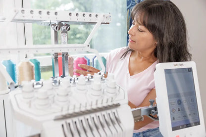 A woman arranges colorful thread spools on a Baby Lock Venture 2 Multi Needle Embroidery Machine by the window, setting up for an embroidery business project. The machine’s digital screen and various threads are visible in front.