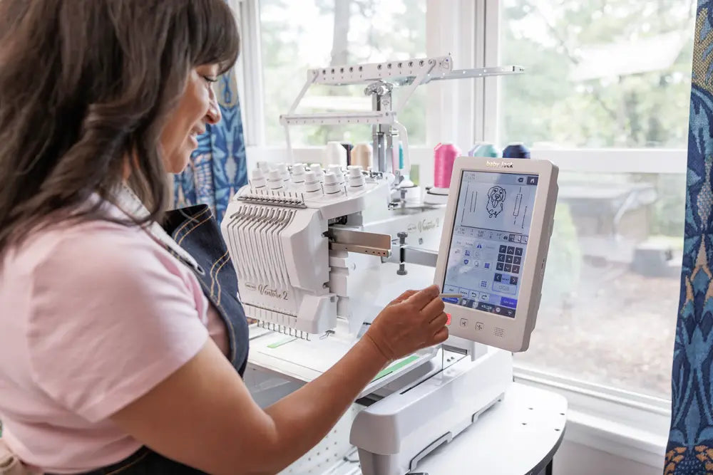 A woman uses the Baby Lock Venture 2 Multi Needle Embroidery Machine, selecting designs on its touchscreen. Colorful threads and natural light from a window brighten her embroidery workspace, showcasing the quality of Baby Lock products.