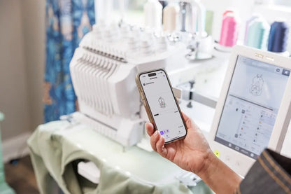 A person holds a smartphone with an embroidery design beside a Baby Lock Venture 2 Multi Needle Embroidery Machine by Baby Lock, showing the same pattern on its touchscreen. Vibrant spools of thread are in the background, ideal for embroidery businesses.
