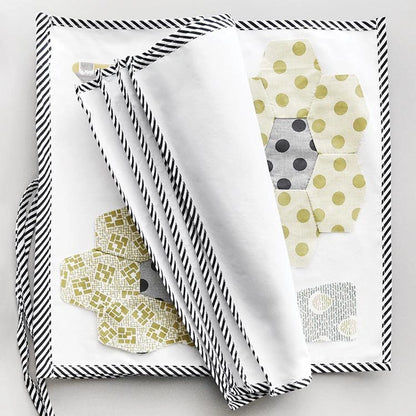 Jen’s Block Wrap by Jen Kingwell Designs is a folded, white organizer with black-and-white striped edges and tie, featuring yellow and gray hexagonal patchwork flower patterns.