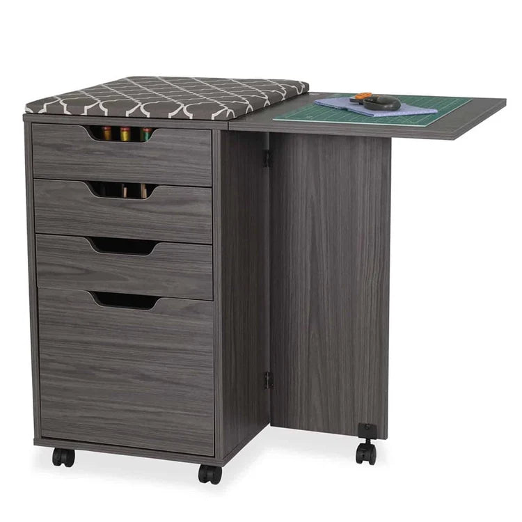 The gray Kiwi Multi-Use Cabinet has four drawers, a cushioned seat, and an extended side table, providing a spacious cutting area for crafting. Sewing tools and a cutting mat are neatly organized on the open table.