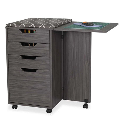 The gray Kiwi Multi-Use Cabinet has four drawers, a cushioned seat, and an extended side table, providing a spacious cutting area for crafting. Sewing tools and a cutting mat are neatly organized on the open table.