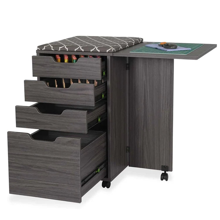 The Kiwi Multi-Use Cabinet is a mobile, gray wood-textured craft station with three open supply drawers, a fold-out cutting surface extension, cushioned seat, and wheels for easy movement.