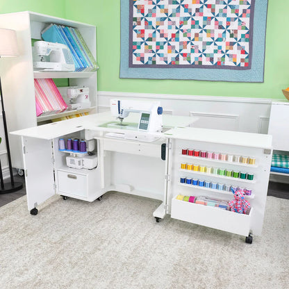 A bright sewing room showcases the Quilted Joy Kangaroo Wallaby Sewing Cabinet Set (available in 2 colors) with a Dual Rail Electric Lift, neatly storing a sewing machine, thread spools, fabric, and tools. A quilt decorates the green wall next to fabric shelves.