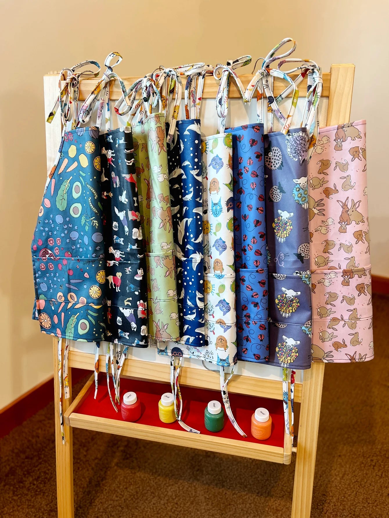 Collection of colorful aprons hanging on a rack with paint bottles below.