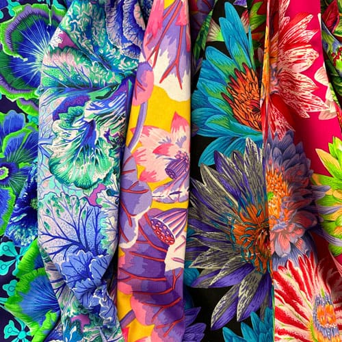 Close-up of vibrant, multicolored fabric featuring bold, large floral patterns in shades of blue, purple, pink, green, yellow, and red, with intricate details and overlapping petal designs.