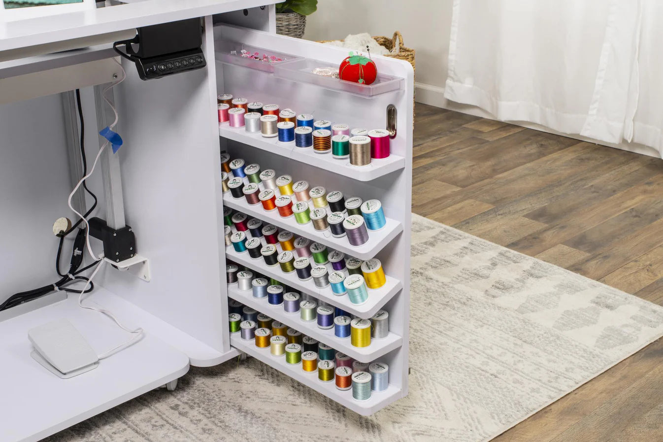 The Koala All-In One Embroidery Studio in White and Gray features organized shelves for colorful thread and a red pin cushion atop the side door. It sits on a wooden floor with a patterned rug nearby, ideal for quilting storage.