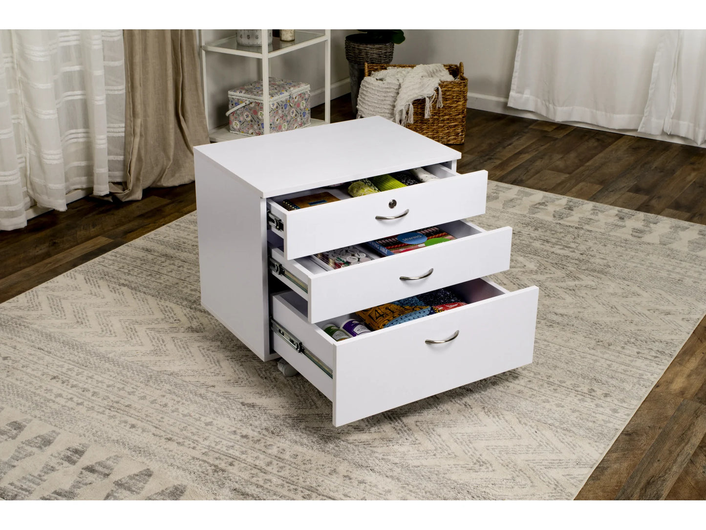 The Koala Quilters Vision Sewing and Companion Chest in White and Gray, with all three drawers open and filled with craft supplies, sits on a patterned rug in a bright room with wooden floors and baskets in the background.