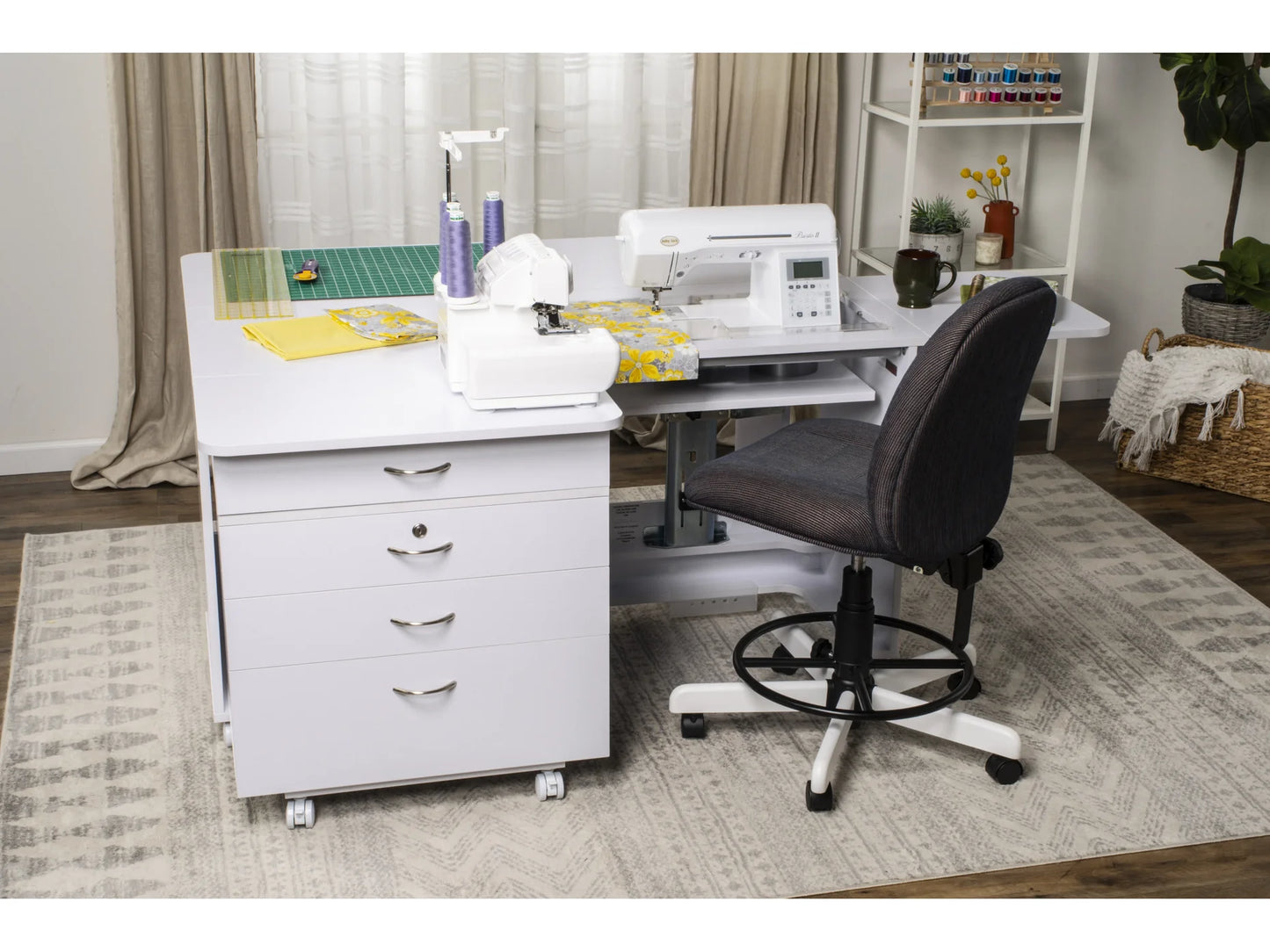 The Koala Quilters Vision Sewing and Companion Chest in White and Gray holds a sewing machine, colorful thread, and fabric on top. A gray chair sits by the chest, with shelves of sewing supplies behind it on a patterned rug.