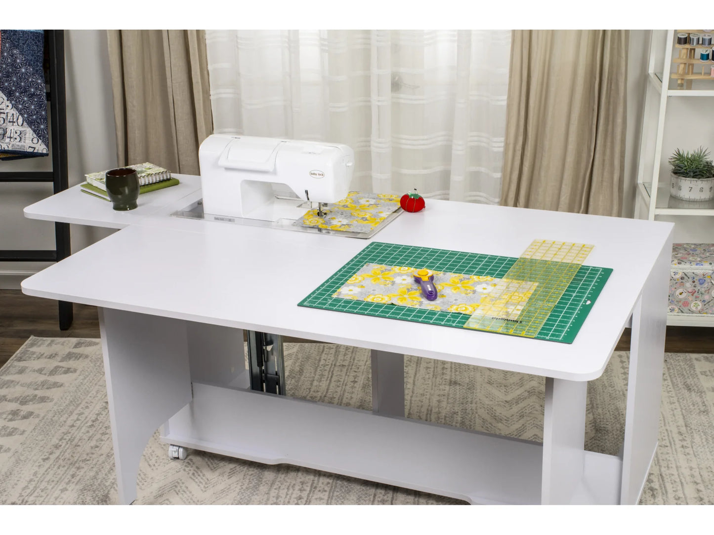 The Koala Quilters Vision Sewing and Companion Chest in White and Gray holds a sewing machine, green cutting mat, rotary cutter, ruler, fabric pieces, red pincushion, and mug in a bright room with curtains and shelves.