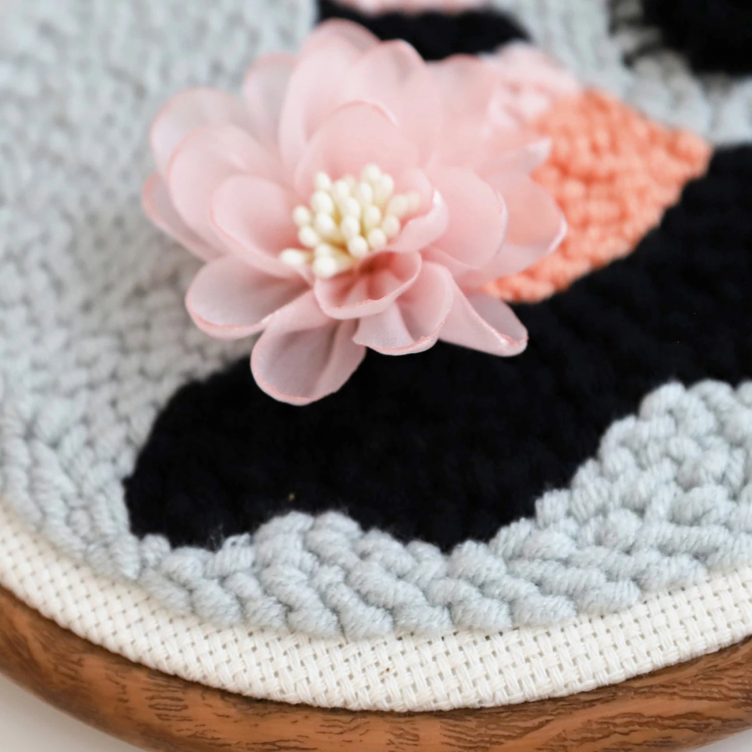 A close-up of a pale pink artificial flower rests on textured, handwoven fabric—ideal for your next Ghouls Night Out Punch Needle Kit project—in shades of gray, black, and peach, with a wooden frame peeking at the edge.