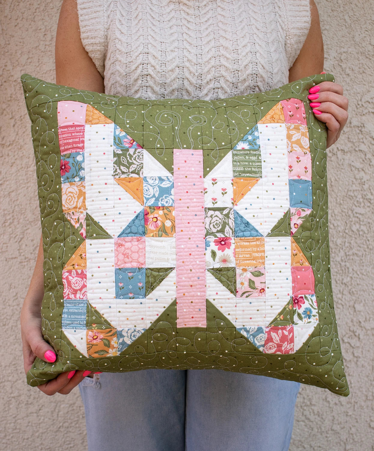 A person in a cream sweater holds a green pillow made with the Mini Charmed Quilt Pattern, featuring vibrant butterflies from assorted floral and geometric fabrics. Their nails are painted pink, highlighting this fun way to use your favorite mini charm pack.