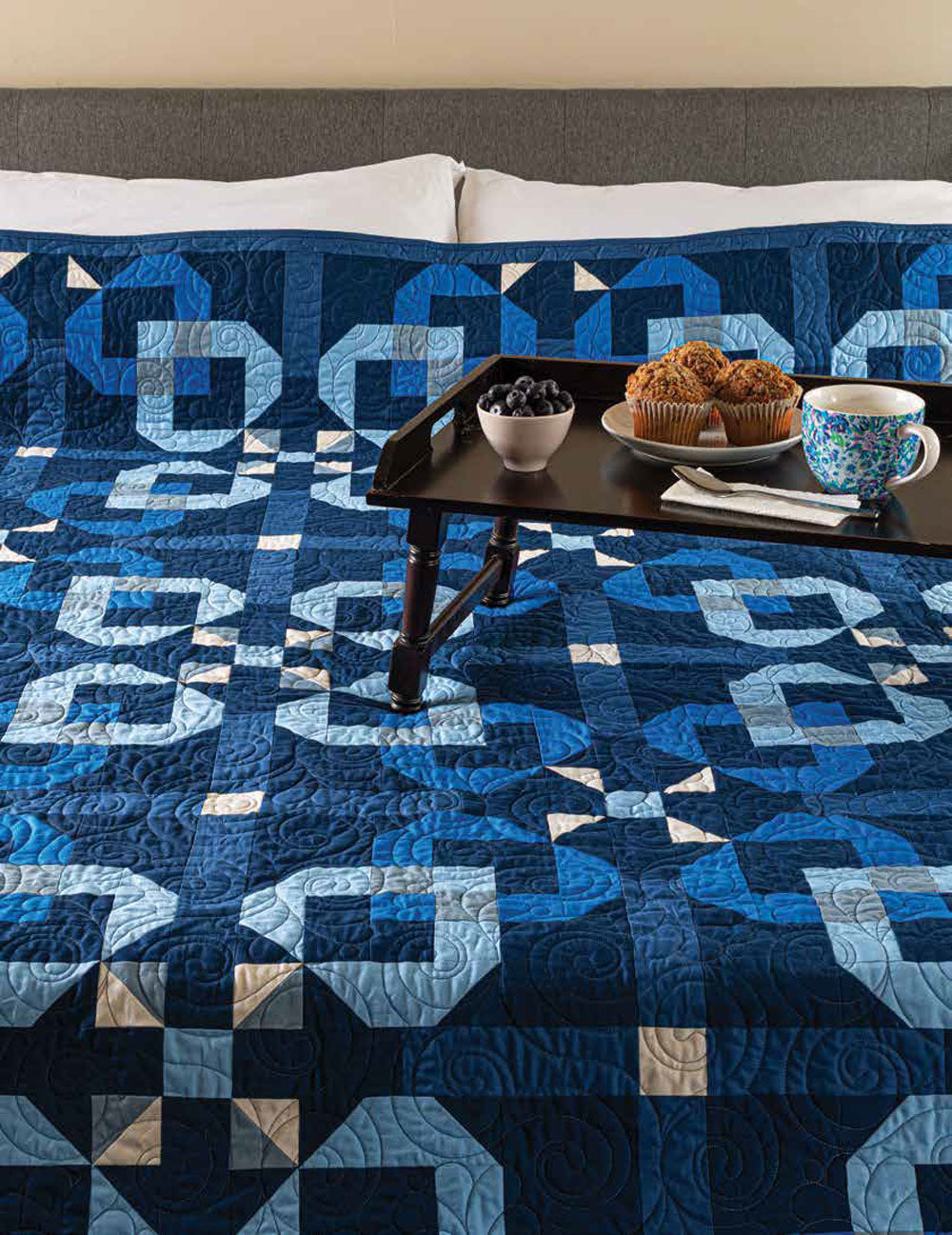 A dark and light blue geometric vintage-style quilt from the Quilted Joy Love of Quilting Blueberry Jam Quilt Kit covers the bed. On top, a black tray with coffee, blueberries, and muffins creates an inviting, cozy scene.
