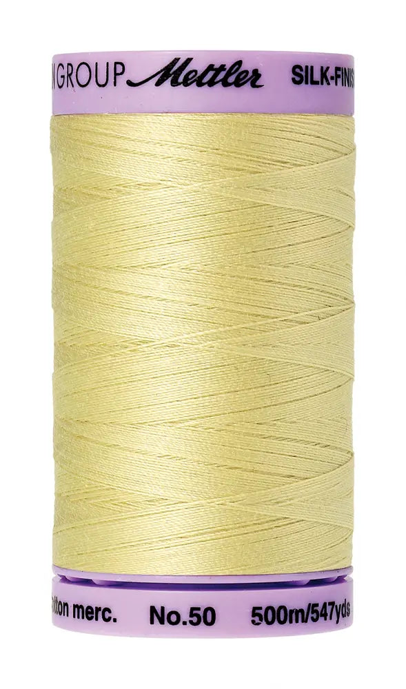A spool of Mettler Cotton Silk Finish 50wt 500m sewing thread in Lemon Frost, with a pale yellow color and pale pink label.