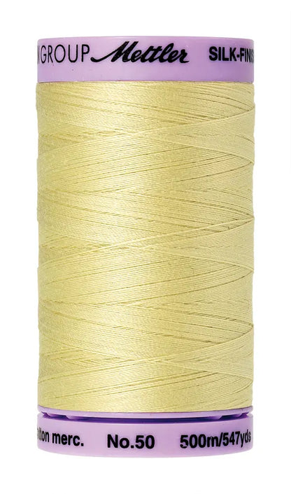 A spool of Mettler Cotton Silk Finish 50wt 500m sewing thread in Lemon Frost, with a pale yellow color and pale pink label.