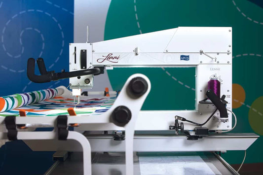 A close-up of the APQS Lenni 22" Quilting Machine with Standard Bliss Table/Frame shows a colorful quilt in progress. The APQS machine features a white body, various controls, and a vibrant blue and green background.