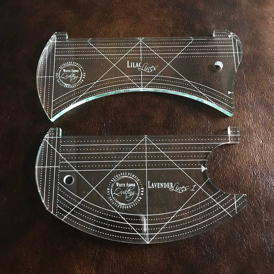 The White Arbor Quilting Lilac & Lavender Duo Ruler Set includes two clear acrylic templates with white grids, circular holes, and engraved branding—perfect as continuous curve rulers on dark brown textured surfaces.