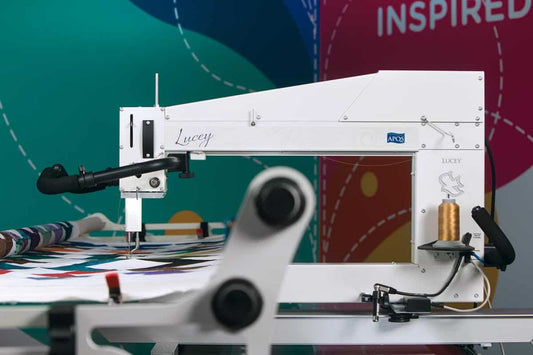 An APQS Demo Lucey 26" Long Arm Quilting Machine by APQS is stitching a colorful quilt, with a gold thread spool on the side and a multicolored backdrop showing the partially visible word "INSPIRED.