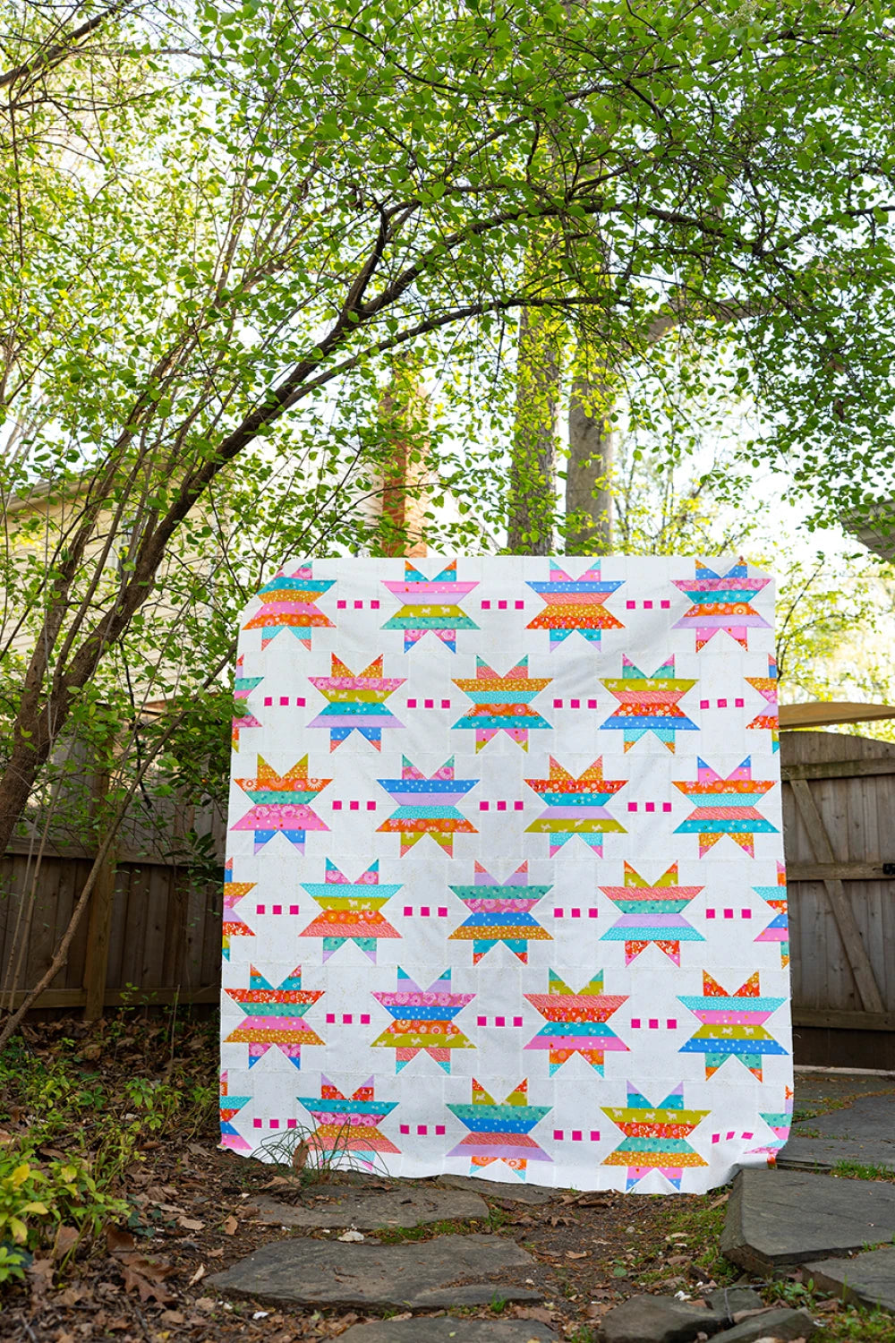 The Spaced Out Quilt Pattern, featuring vibrant galaxies, star motifs, and pink squares on a white background, is displayed outdoors draped over a wooden fence with lush green trees in the backdrop.
