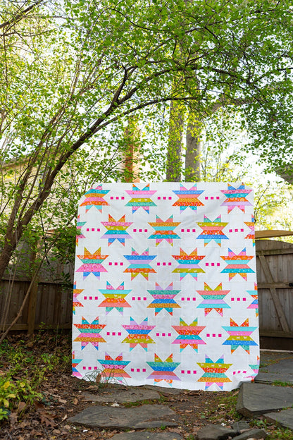 The Spaced Out Quilt Pattern, featuring vibrant galaxies, star motifs, and pink squares on a white background, is displayed outdoors draped over a wooden fence with lush green trees in the backdrop.