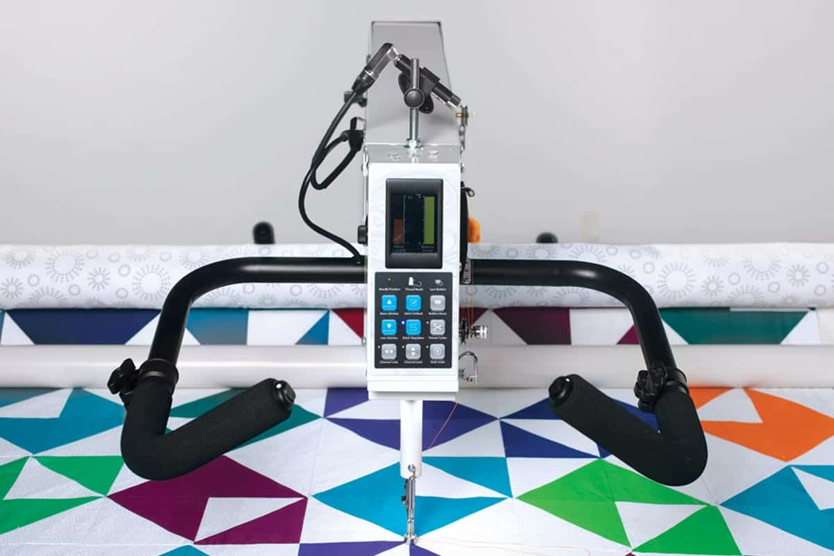 An APQS Demo Millie 30" Long Arm Quilting Machine with handlebars and touchscreen is set over a vibrant geometric quilt featuring blue, green, orange, and purple patterns.