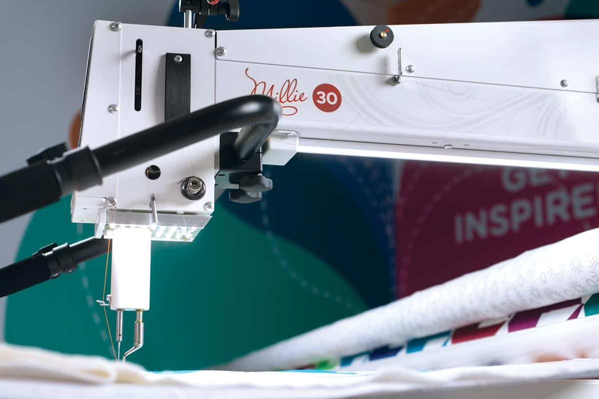 A close-up of the APQS Demo Millie 30" Long Arm Quilting Machine in action, featuring the APQS brand label, light, and needle as it stitches a colorful quilt.