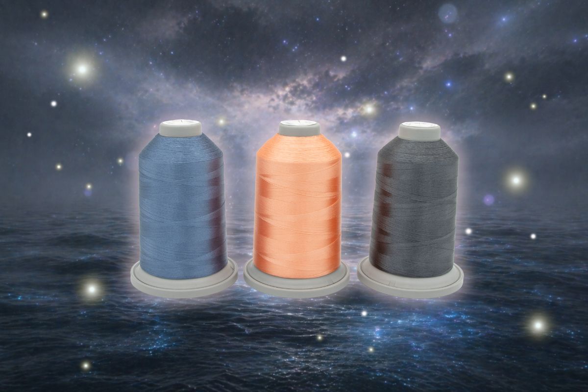 The Moonlight Tides Glide Thread of the Month Bundle by Hab+Dash by Fil Tec features three large spools—blue, peach, and dark gray—displayed on a water-like surface with a starry galaxy backdrop, ideal for quilting or embroidery.
